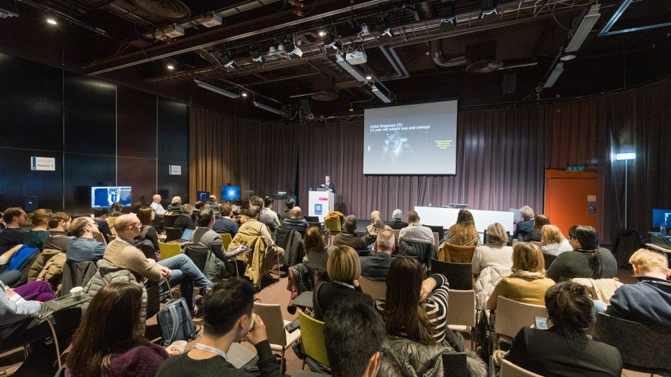 A man and a woman stand by the podium in discussion. Behind them on the screen you can see the Karolinska University Hospital and the Karolinska Institutet.