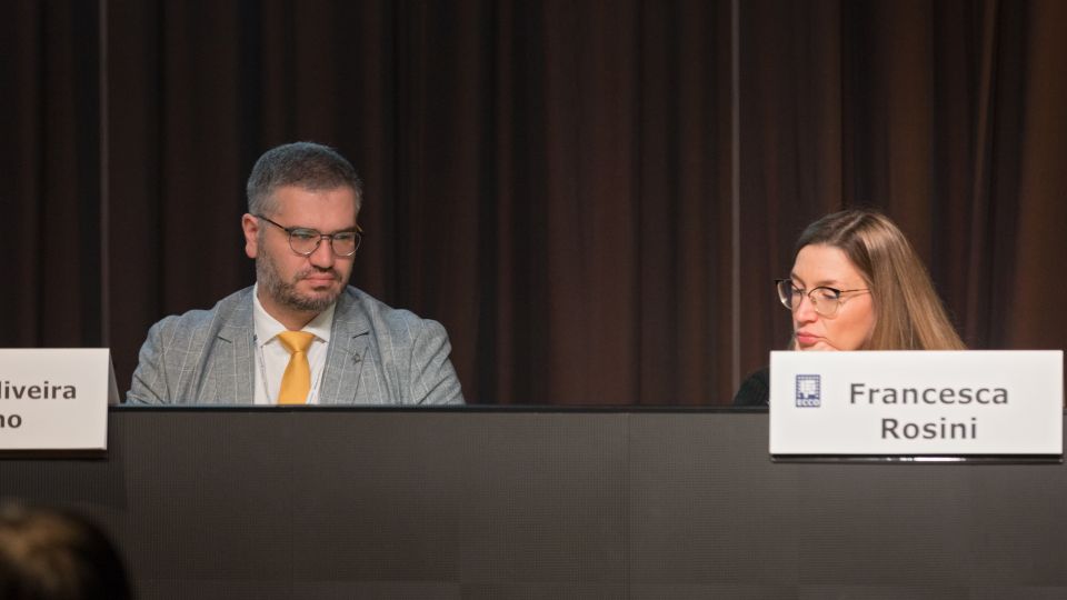 A man and a woman stand by the podium in discussion. Behind them on the screen you can see the Karolinska University Hospital and the Karolinska Institutet.
