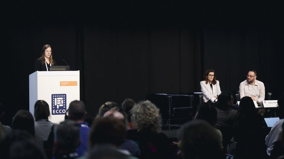 A man and a woman stand by the podium in discussion. Behind them on the screen you can see the Karolinska University Hospital and the Karolinska Institutet.