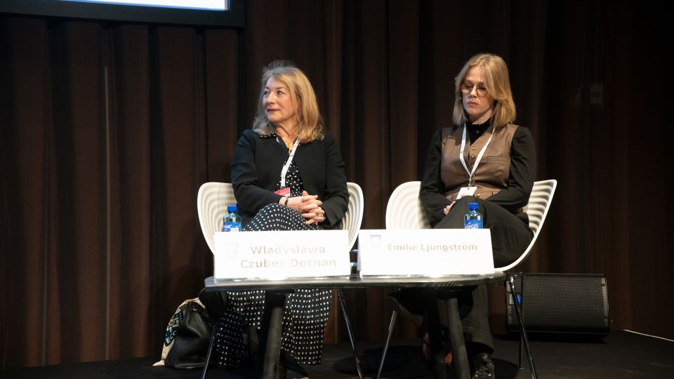 Marc Ferrante and Konstantinos Papamichail sit next to the podium behind a long desk. In the background you can see the speakers conversing and the screen with a graphic of the Swedish Inflamatory Bowel Disease Registry.