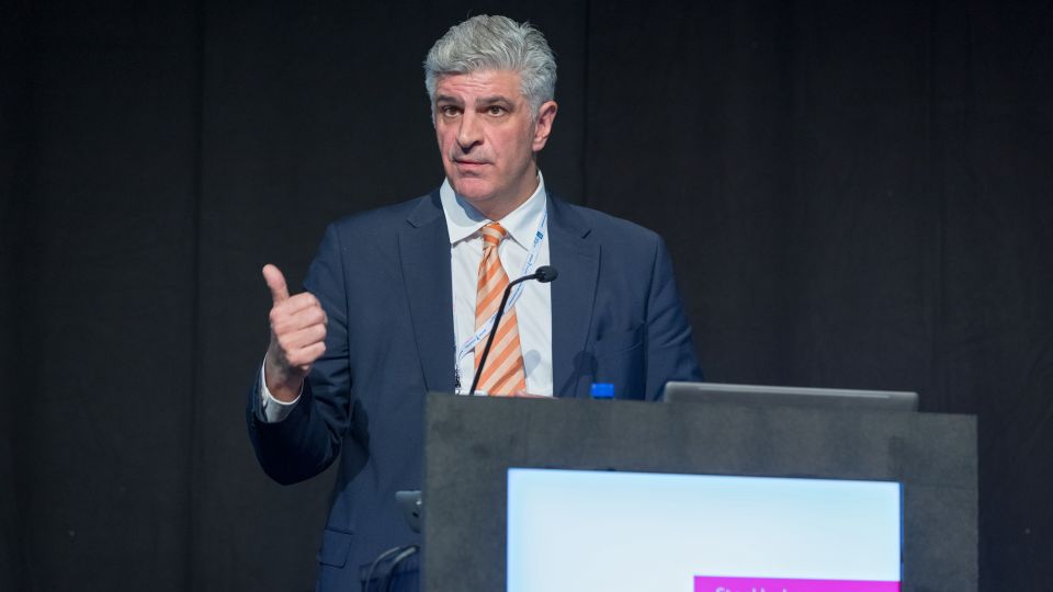 Marc Ferrante and Konstantinos Papamichail sit next to the podium behind a long desk. In the background you can see the speakers conversing and the screen with a graphic of the Swedish Inflamatory Bowel Disease Registry.