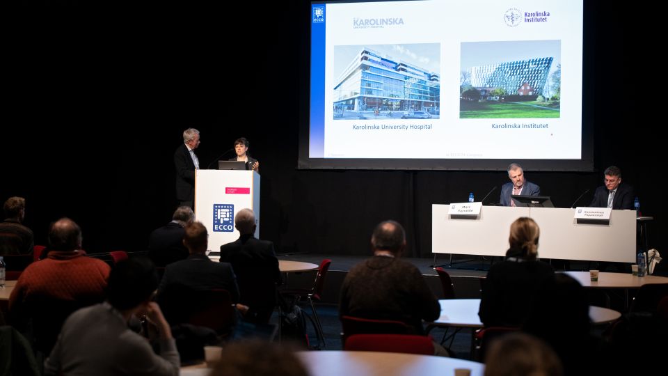 A man and a woman stand by the podium in discussion. Behind them on the screen you can see the Karolinska University Hospital and the Karolinska Institutet.