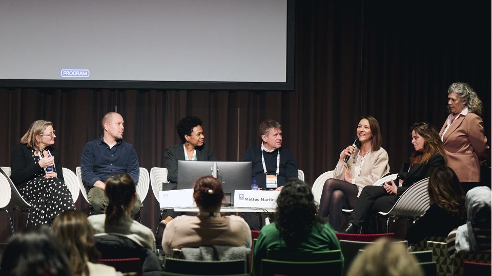 Marc Ferrante and Konstantinos Papamichail sit next to the podium behind a long desk. In the background you can see the speakers conversing and the screen with a graphic of the Swedish Inflamatory Bowel Disease Registry.