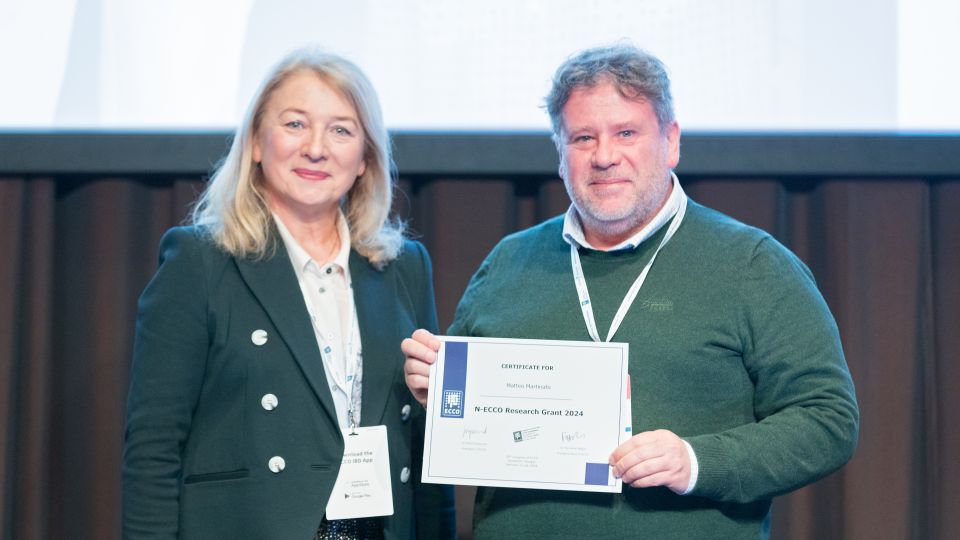 Marc Ferrante and Konstantinos Papamichail sit next to the podium behind a long desk. In the background you can see the speakers conversing and the screen with a graphic of the Swedish Inflamatory Bowel Disease Registry.