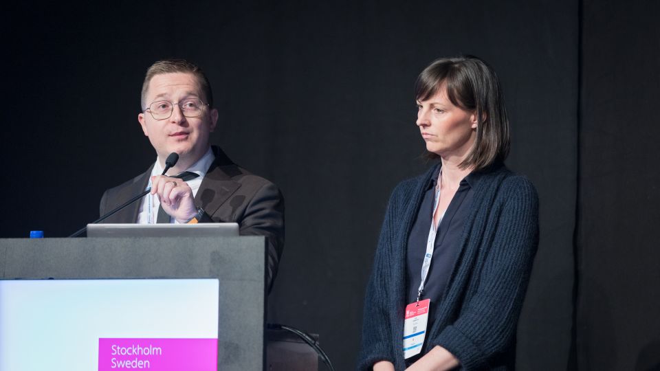 A man and a woman stand by the podium in discussion. Behind them on the screen you can see the Karolinska University Hospital and the Karolinska Institutet.