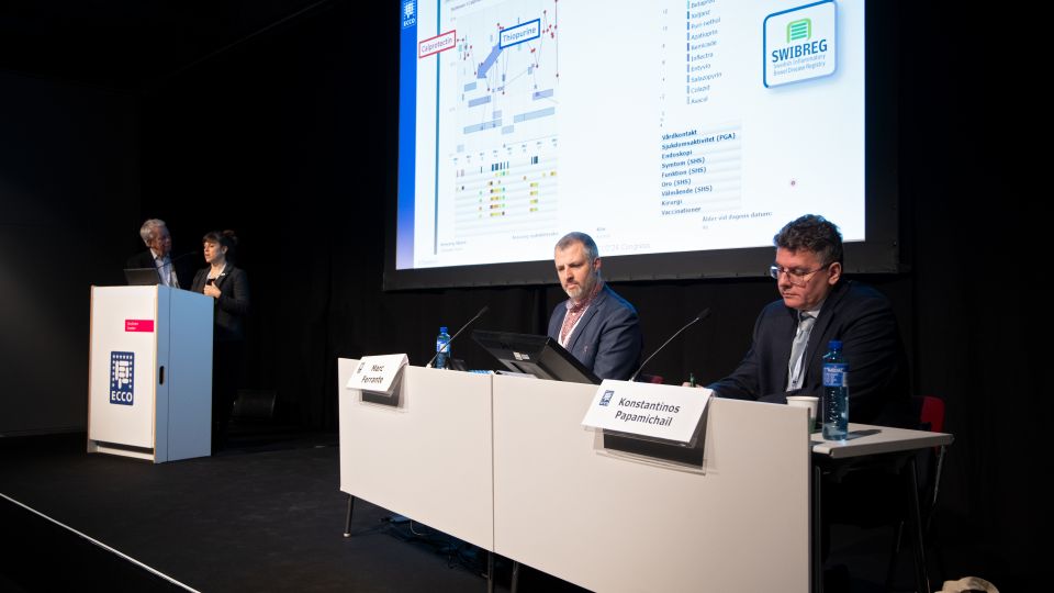 Marc Ferrante and Konstantinos Papamichail sit next to the podium behind a long desk. In the background you can see the speakers conversing and the screen with a graphic of the Swedish Inflamatory Bowel Disease Registry.