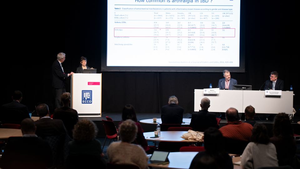 On the screen behind the speakers and the experts is a table showing how common arthralgia is in IBD