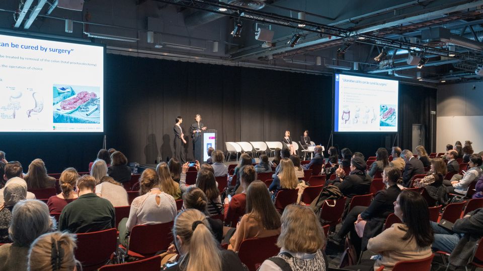 Marc Ferrante and Konstantinos Papamichail sit next to the podium behind a long desk. In the background you can see the speakers conversing and the screen with a graphic of the Swedish Inflamatory Bowel Disease Registry.