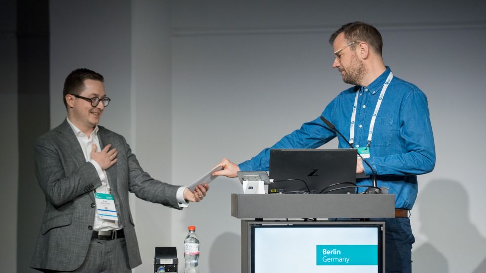 Marc Ferrante and Konstantinos Papamichail sit next to the podium behind a long desk. In the background you can see the speakers conversing and the screen with a graphic of the Swedish Inflamatory Bowel Disease Registry.