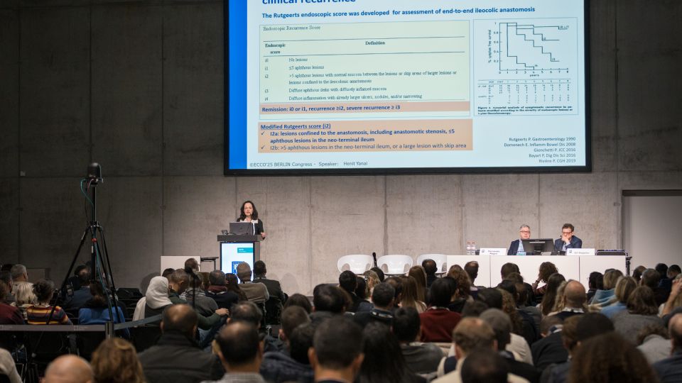 Marc Ferrante and Konstantinos Papamichail sit next to the podium behind a long desk. In the background you can see the speakers conversing and the screen with a graphic of the Swedish Inflamatory Bowel Disease Registry.