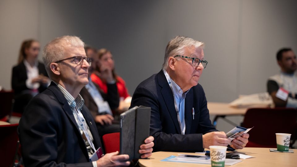 Two male participants, dressed formally in suits, follow the talk with concentration and cups of coffees.