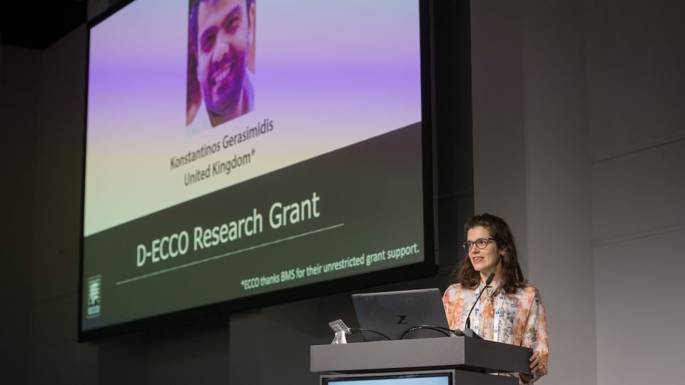 Marc Ferrante and Konstantinos Papamichail sit next to the podium behind a long desk. In the background you can see the speakers conversing and the screen with a graphic of the Swedish Inflamatory Bowel Disease Registry.