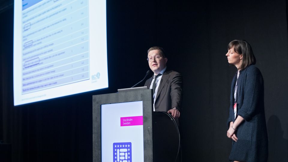 Marc Ferrante and Konstantinos Papamichail sit next to the podium behind a long desk. In the background you can see the speakers conversing and the screen with a graphic of the Swedish Inflamatory Bowel Disease Registry.