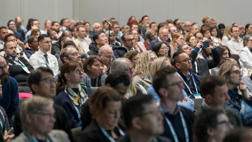 Two male participants, dressed formally in suits, follow the talk with concentration and cups of coffees.
