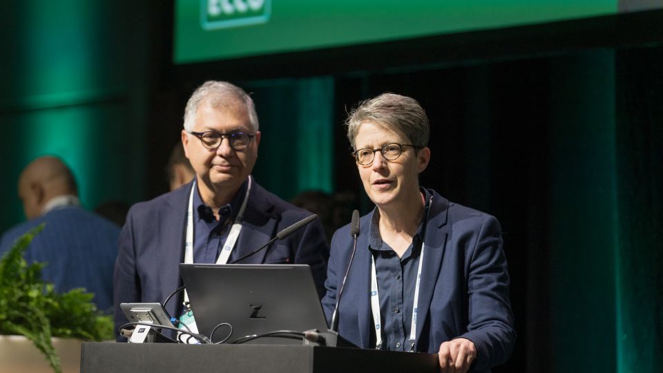 Marc Ferrante and Konstantinos Papamichail sit next to the podium behind a long desk. In the background you can see the speakers conversing and the screen with a graphic of the Swedish Inflamatory Bowel Disease Registry.