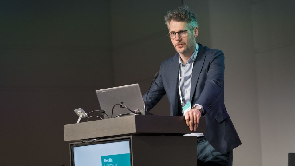 Marc Ferrante and Konstantinos Papamichail sit next to the podium behind a long desk. In the background you can see the speakers conversing and the screen with a graphic of the Swedish Inflamatory Bowel Disease Registry.