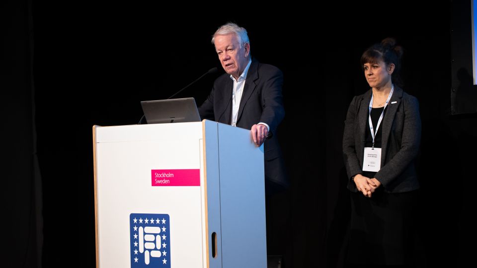 One speaker stands behind the podium, the other next to him.