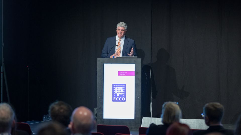 A man and a woman stand by the podium in discussion. Behind them on the screen you can see the Karolinska University Hospital and the Karolinska Institutet.