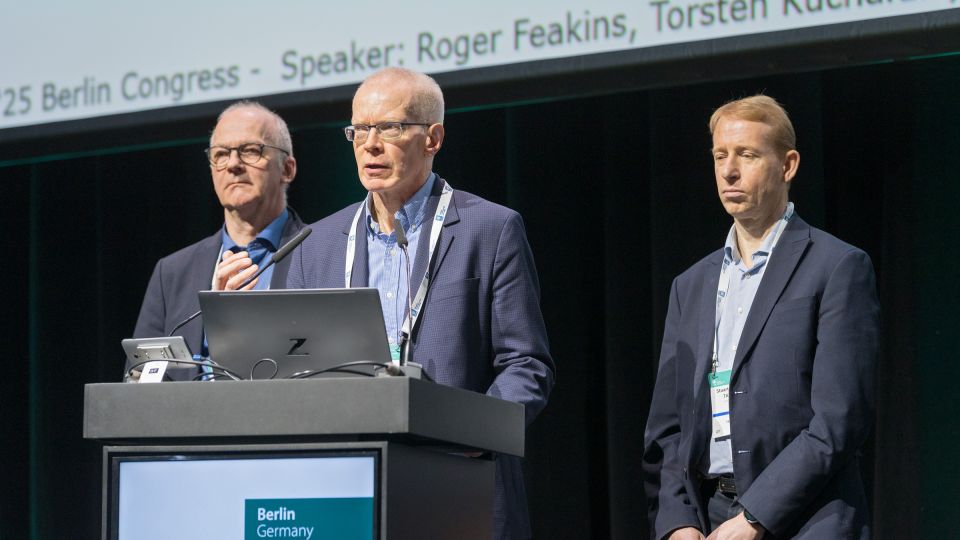 Marc Ferrante and Konstantinos Papamichail sit next to the podium behind a long desk. In the background you can see the speakers conversing and the screen with a graphic of the Swedish Inflamatory Bowel Disease Registry.