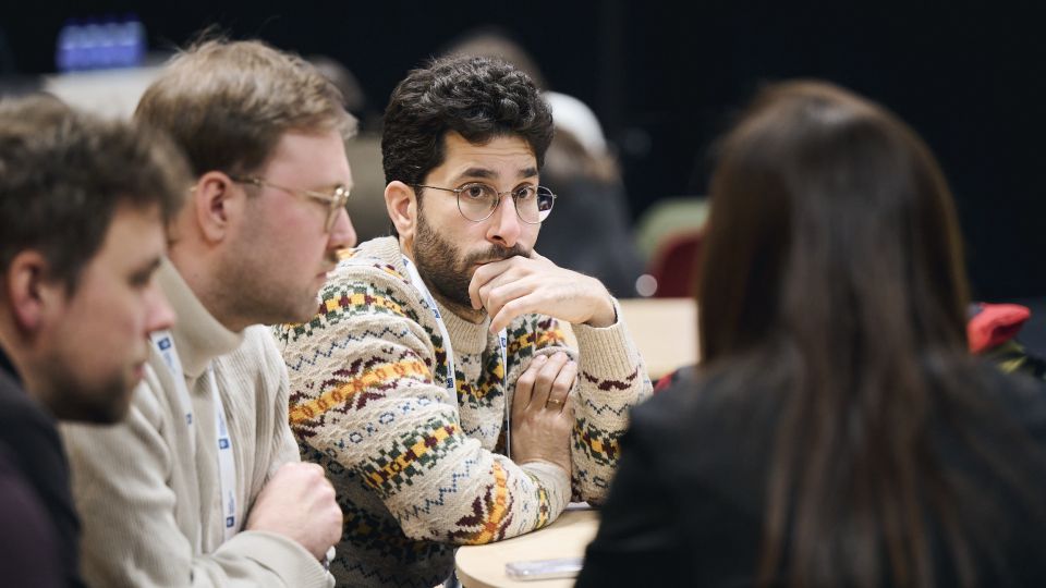 Two male participants, dressed formally in suits, follow the talk with concentration and cups of coffees.
