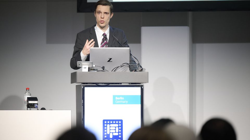 A man and a woman stand by the podium in discussion. Behind them on the screen you can see the Karolinska University Hospital and the Karolinska Institutet.