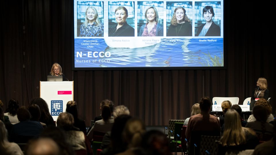 A man and a woman stand by the podium in discussion. Behind them on the screen you can see the Karolinska University Hospital and the Karolinska Institutet.