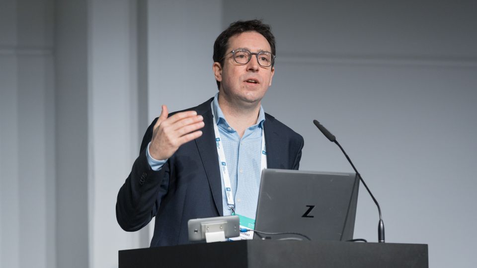 Marc Ferrante and Konstantinos Papamichail sit next to the podium behind a long desk. In the background you can see the speakers conversing and the screen with a graphic of the Swedish Inflamatory Bowel Disease Registry.
