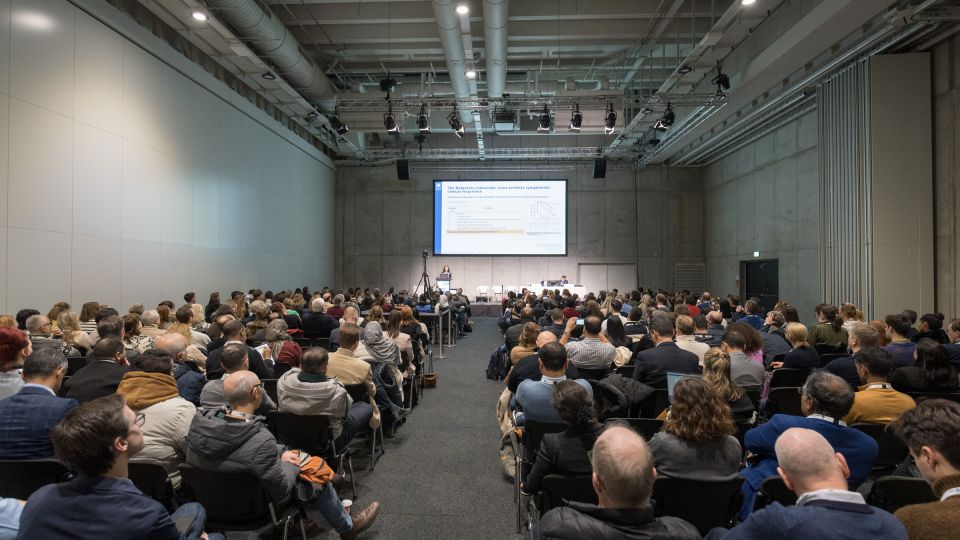 A man and a woman stand by the podium in discussion. Behind them on the screen you can see the Karolinska University Hospital and the Karolinska Institutet.