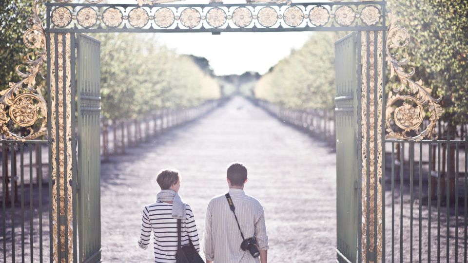 Two people with bag and camera walk through an golden ornamented park gate.