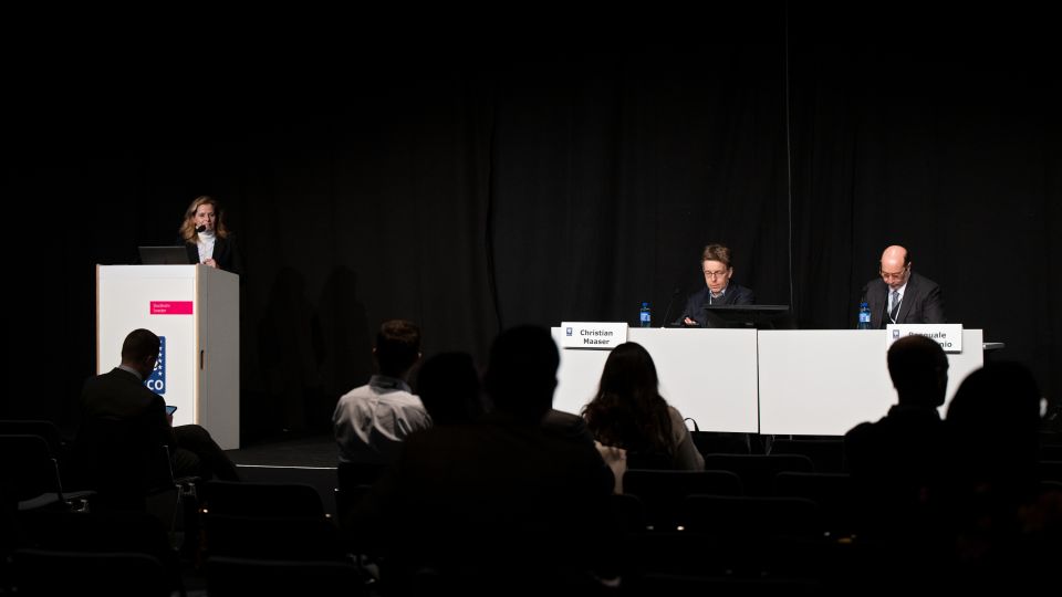 Marc Ferrante and Konstantinos Papamichail sit next to the podium behind a long desk. In the background you can see the speakers conversing and the screen with a graphic of the Swedish Inflamatory Bowel Disease Registry.