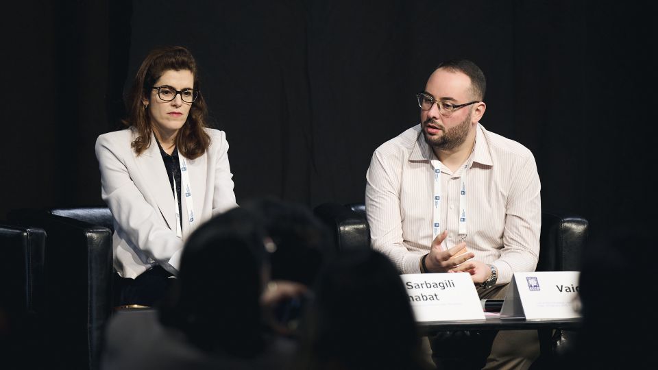 Marc Ferrante and Konstantinos Papamichail sit next to the podium behind a long desk. In the background you can see the speakers conversing and the screen with a graphic of the Swedish Inflamatory Bowel Disease Registry.