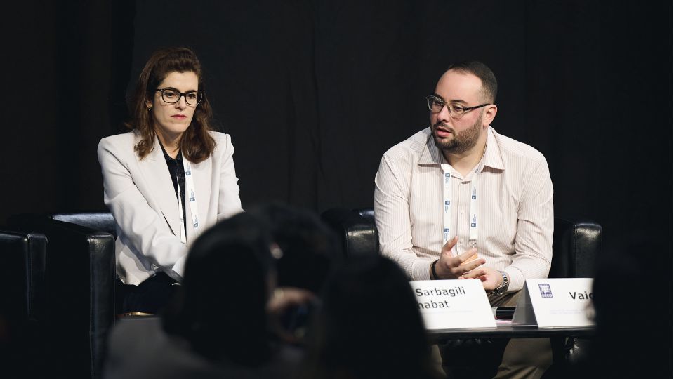 Marc Ferrante and Konstantinos Papamichail sit next to the podium behind a long desk. In the background you can see the speakers conversing and the screen with a graphic of the Swedish Inflamatory Bowel Disease Registry.