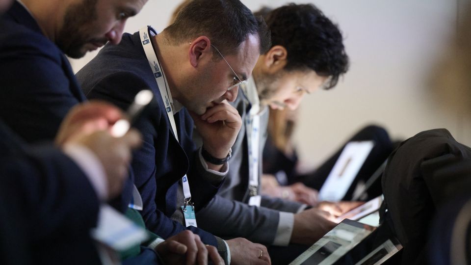 Two male participants, dressed formally in suits, follow the talk with concentration and cups of coffees.