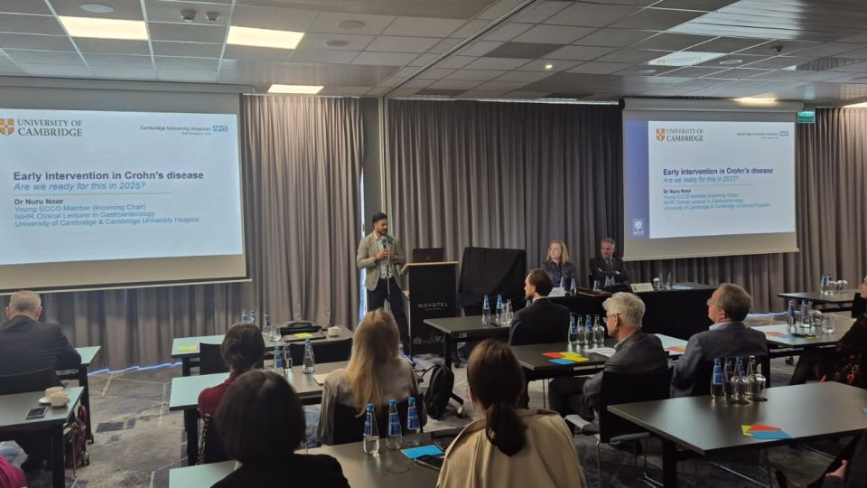 Marc Ferrante and Konstantinos Papamichail sit next to the podium behind a long desk. In the background you can see the speakers conversing and the screen with a graphic of the Swedish Inflamatory Bowel Disease Registry.