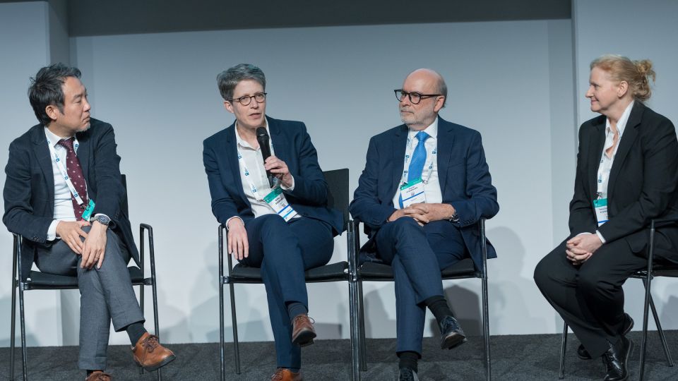 Marc Ferrante and Konstantinos Papamichail sit next to the podium behind a long desk. In the background you can see the speakers conversing and the screen with a graphic of the Swedish Inflamatory Bowel Disease Registry.