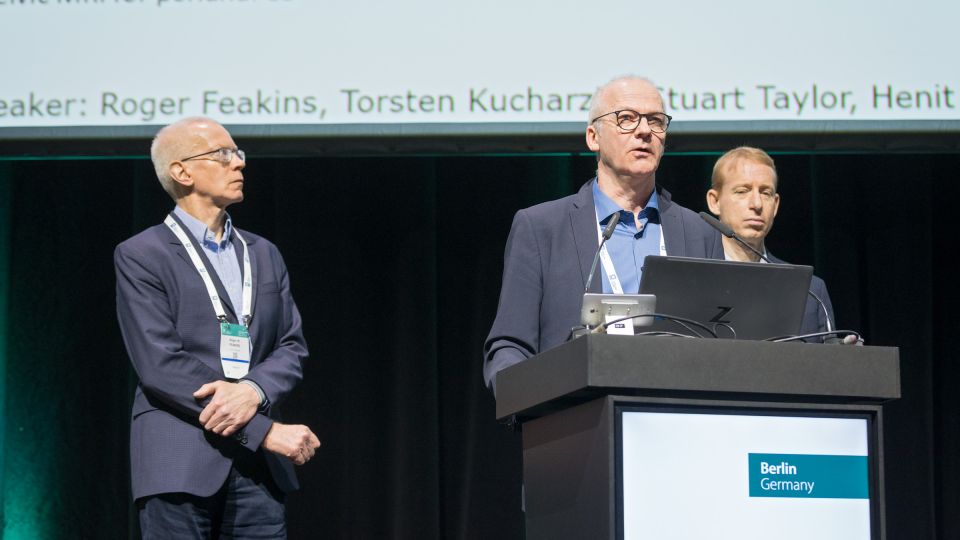 A man and a woman stand by the podium in discussion. Behind them on the screen you can see the Karolinska University Hospital and the Karolinska Institutet.