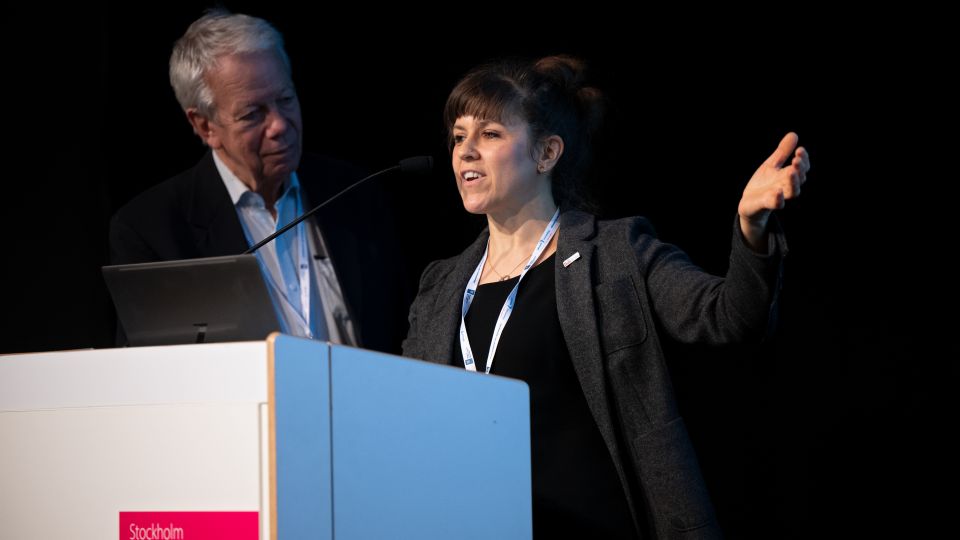The female speaker talks and gestures enthusiastically behind the podium